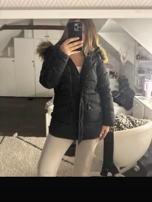 Svart lång dunjacka med fuskpäls - Snygg svart dunjacka med huva och fuskpälsdetalj. Jackan är lång, har dragkedja och knappar framtill samt stora fickor med lock. Perfekt för kalla vinterdagar och ger en riktigt stilren look.