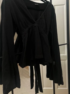 Svart v-ringad topp med knyte - Snygg svart omlottopp med v-ringning och långa ärmar. Toppen har knytband i midjan och ett lätt utställt fall nedtill. Perfekt för dig som gillar en stilren och trendig look.