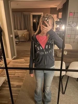 Mörkblå zip hoodie med tryck - Mörkblå hoodie med dragkedja och vit text på bröstet. Modellen har huva med vita snören och fickor framtill. Perfekt för en avslappnad och cool stil. Passar till jeans eller mjukisbyxor.