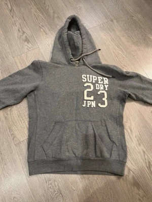 Grå hoodie Superdry med tryck - Säljer en grå hoodie från Superdry med vitt tryck på bröstet där det står 'SUPER DRY 23 JPN'. Hoodien har huva med dragsko och en stor magficka. Perfekt för chill dagar och streetwear-stil.