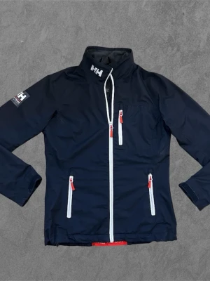 Marinblå vindjacka från Helly Hansen - Snygg jacka från Helly Hansen, passar perfekt för blåsiga dagar och inför hösten. Texten där bak har lossat lite. Den har mycket kvar att ge. Strl: S/XS 