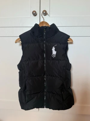 Svart dunväst från Polo Ralph Lauren - Snygg svart dunväst från Polo Ralph Lauren med hög krage och dragkedja framtill. Västen har ett stort vitt Polo-logotryck på bröstet och två sidofickor. Har även en innerficka. Perfekt för lager-på-lager och håller dig varm under kyliga dagar.