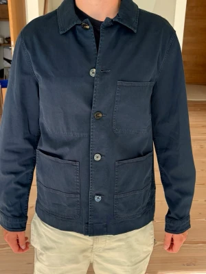 Mörkblå overshirt från woolrich - Helt ny Stilren mörkblå overshirt från Woolrich i bomull med tre stora utanpåliggande fickor och knäppning framtill. Lapparna finns kvar. Klassisk krage och raka ärmar ger en clean look. Perfekt att bära över en t-shirt för en avslappnad men snygg stil. Nypris 2800 kr