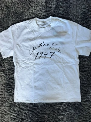 Vit t-shirt med Christian Dior-tryck - Vit t-shirt med svart text 'Christian Dior 1947' på bröstet. Klassisk rund hals och korta ärmar. T-shirten har en enkel och stilren design som passar till det mesta. Tillverkad i mjukt bomullsmaterial för skön känsla.