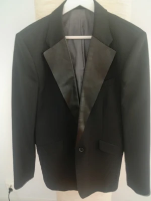 Svart smokingkavaj från Acne.  - Snygg svart smokingkavaj från Acne med klassiska slag i blankt läder. Kavajen har en enkelknäppning och innerfoder i grått. Perfekt för dig som vill ha en stilren och modern look. Strl.) 50. Minimal slitning på ena axeln. Svårt att få en vettig bild på, då den är ytterst liten. 