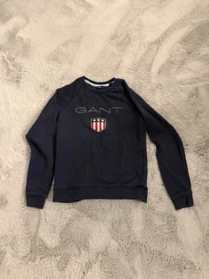Mörkblå sweatshirt från GANT - Mörkblå sweatshirt från GANT med broderad logga och ett sköldmärke med stjärnor och ränder på bröstet. Klassisk rund halsringning och ribbade muddar vid ärmslut och nederkant. Perfekt för en avslappnad och stilren look.