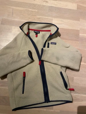 Beige fleecejacka från Patagonia - Mysig beige fleecejacka från Patagonia med marinblå detaljer och dragkedjor. Jackan har hög krage, två fickor med dragkedja och röda detaljer på dragkedjorna. Perfekt för lager-på-lager och outdoor-vibes.