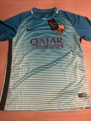 Barcelona Neymar Jr matchtröja Nike - Snygg Barcelona matchtröja från Nike med Neymar Jr och nummer 11 på ryggen. Tröjan är turkos med mörkblå ärmar och tunna horisontella ränder, klubbmärke och sponsortryck på bröstet. Perfekt för fotbollsträning eller att bära på läktaren.
