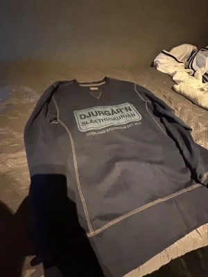Mörkblå sweatshirt Djurgår'n - Mörkblå sweatshirt med tryck på bröstet där det står 'DJURGÅR'N SLAKTHUSKURVAN DFFENDERS STOCKHOLM EST. 2015'. Tröjan har kontrastsömmar och rund hals. Perfekt för dig som vill visa ditt stöd för Djurgården.