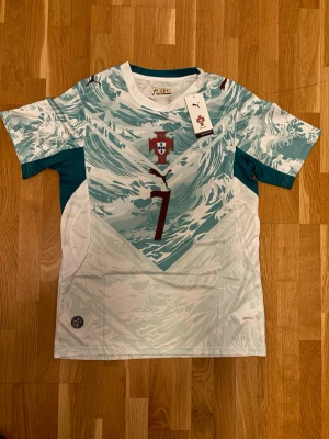 Portugal fotbollströja Ronaldo Puma - Portugal fotbollströja med Ronaldo 7-tryck, tillverkad av Puma. Tröjan har ett unikt mönster i ljusblått och vitt, med Portugals landslagsmärke på bröstet och Puma-logga. Korta ärmar och sportig passform, perfekt för fotboll eller träning.
