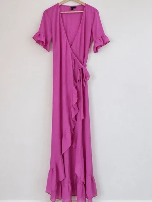 Lång rosa omlottklänning med volang - Snygg lång rosa omlottklänning med volangdetaljer längs kanten och nederdelen. Klänningen har korta ärmar med volang och knyt i midjan för en feminin siluett. Perfekt för sommarens alla tillfällen.