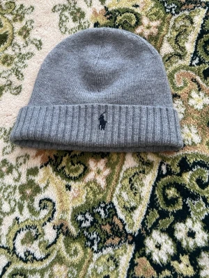 Grå ribbstickad mössa Polo Ralph Lauren - Snygg grå mössa från Polo Ralph Lauren med ribbstickad kant och den klassiska broderade loggan framtill. Tillverkad i mjukt stickat material som håller dig varm och stilren under kalla dagar.