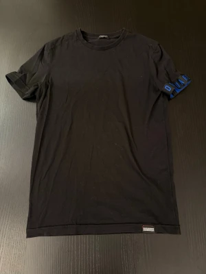 D-squared2 Svart t-shirt blå  - Svart t-shirt med korta ärmar och blå D-squared2 på ena ärmen. Tillverkad i mjuk bomull och har en klassisk passform. Liten etikett nertill med D-squared2 Perfekt till vardags för en sportig look.