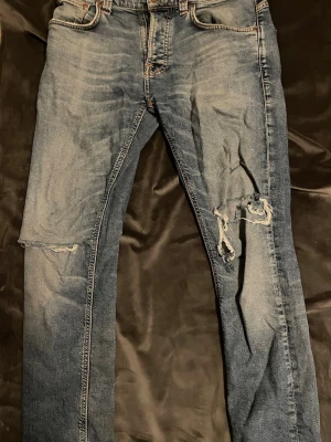 Nudie jeans  - Ett par blå jeans i slim fit-modell med slitningar och stora hål på höger ben. Jeansen har klassisk femficksdesign, ljus tvätt och detaljerade sömmar. Perfekta för en avslappnad och trendig look.