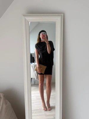 Draped Mini Cape Dress - Snygg mörkbrun kortklänning med draperad överdel och ärmlös design. Klänningen har en loose passform upptill och smalare kjol, vilket ger en cool och modern siluett. Perfekt för dig som gillar minimalistisk stil och vill sticka ut med enkla detaljer.