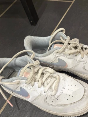 Vita Nike sneakers med pastell swoosh - Säljer ett par vita Nike sneakers med lågt skaft. Skorna har en snygg swoosh i ljusblått och ljusrosa på sidan, samt vita snören och grått innerfoder. Ovandelen är i läder med perforeringar på tån för extra ventilation. Perfekta för en clean och stilren look.