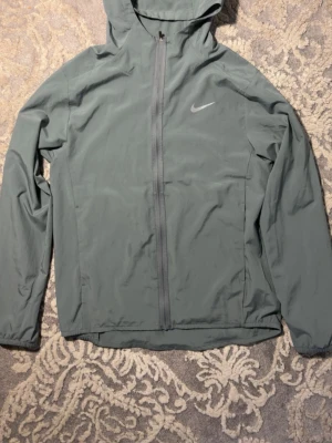 Grå vindjacka med huva från Nike - Snygg grå vindjacka från Nike med hel dragkedja framtill och huva. Jackan har diskret Nike-logga på bröstet, långa ärmar och elastiska muddar. Tillverkad i lätt och smidigt material som passar perfekt för blåsiga dagar. Pris kan diskuteras!