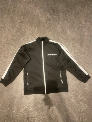 Palm Angels - Snygg svart track jacket från Palm Angels med vita ränder längs ärmarna och vit dragkedja. Jackan har hög krage, två fickor med dragkedja och logga på bröstet. Perfekt för en sportig och trendig streetwear-look.