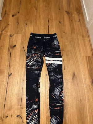 Mönstrade leggings från Stronger - Snygga svarta leggings från Stronger med färgstarkt safari-inspirerat mönster i vitt, orange och grått. Hög midja med logotyp i midjebandet och två vita ränder på ena benet. Perfekta för träning eller chill. Materialet är stretchigt och följsamt.