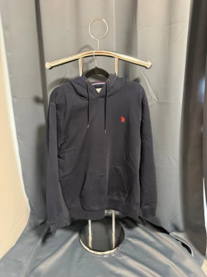 US POLO hoodie  - Mörkblå hoodie från U.S. Polo Assn. med klassisk huva och dragsnören. Tröjan har en känguruficka framtill och en liten röd logga broderad på bröstet. Tillverkad i mjukt material som passar perfekt för en avslappnad stil.