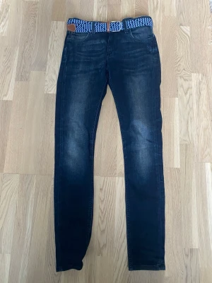 Mörkblå skinny jeans från Lee - Snygga mörkblå skinny jeans från Lee med klassisk femficksmodell och subtila slitningar. Jeansen har en blåvit flätad bälte och är tillverkade i stretchigt denimtyg för extra komfort. Perfekta för en avslappnad och trendig look. plus att man får ett ljus / mörkt blått bälte från onewear.