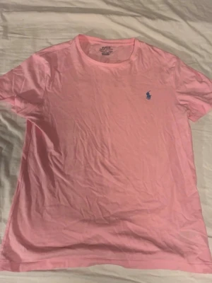 Rosa t-shirt från Ralph Lauren - Snygg rosa t-shirt från Ralph Lauren med klassisk passform och blå logga broderad på bröstet. Tillverkad i mjuk bomull och har rund halsringning samt korta ärmar. Perfekt för en avslappnad och stilren look.