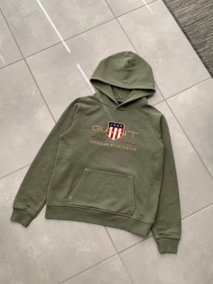 Olivgrön hoodie från GANT - Säljer en olivgrön hoodie från GANT med klassiskt tryck och logga på bröstet. Hoodien har en stor ficka framtill, ribbade muddar och huva. Materialet är mjukt och bekvämt, perfekt för dig som gillar en sportig och avslappnad stil. Storlek 158-164cm - 13-14 år.