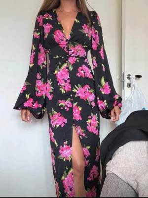 Adoore Positano  - Svart långklänning med stora rosa blommor, v-ringad och med långa, vida ärmar. Klänningen har en slits framtill och markerad midja. Perfekt för dig som vill sticka ut med ett färgstarkt mönster och elegant siluett. Extra unik då detta mönster inte längre säljs på hemsidan.