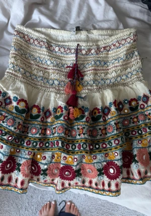 Broderad kortkjol med volang och tofsar - Färgglad kortkjol med broderade blommor i rött, rosa, gult och grönt på vit botten. Kjolen har volang, smock i midjan och dekorativa tofsar framtill. Perfekt för dig som gillar boho-stil och detaljer.
