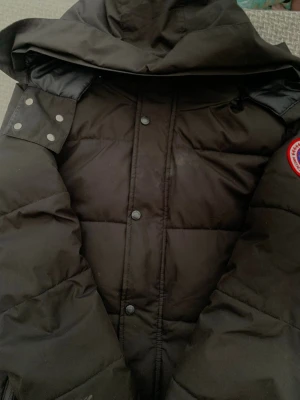Svart dunjacka från Canada Goose - Säljer en svart dunjacka från Canada Goose med huva och klassisk logotyp-patch på ärmen. Jackan har tryckknappar framtill och breda, quiltade paneler som ger en riktigt varm känsla. Perfekt för kalla vinterdagar.