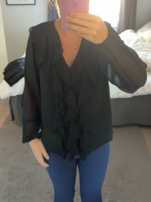 Svart blus med volangdetaljer - Snygg svart blus med långa ärmar i ett tunt, transparent material. Blusen har djup V-ringning och volangdetaljer framtill som ger en trendig look. Perfekt att styla med jeans eller kjol för en chic outfit.