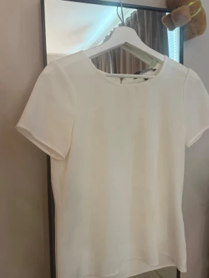 Vit enkel kortärmad topp  - Säljer en stilren vit topp med rund halsringning och korta ärmar. Toppen har en clean look utan mönster eller detaljer och passar perfekt till många olika outfits. Materialet känns mjukt och lätt, troligen polyester eller bomull.