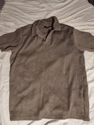 Brun piketröja  - INSTABOX!!! Säljer en brun ribbad skjorta med korta ärmar och klassisk krage. Skjortan har en relaxed fit och är tillverkad i ett mjukt material som ger en chill vibe. Perfekt för dig som gillar stilrena plagg med lite textur. XS herr storlek.