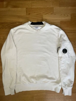 Vit sweatshirt från C.P. Company - Vit sweatshirt från C.P. Company med rund halsringning och ribbade muddar. Tröjan har en ikonisk svart linsdetalj på ena ärmen och är tillverkad i mjuk bomull. Perfekt för en clean och stilren look.