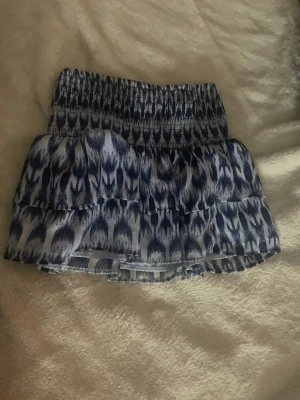 Blåvit kortkjol med volanger - Supersöt kortkjol i blått och vitt med elastisk midja och volangdetaljer. Kjolen har ett mönster som påminner om batik eller abstrakta ränder och är perfekt för varma dagar. Lätt och luftig känsla, passar till både sneakers och sandaler.