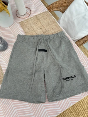 Gråa Essentials shorts med snörning - Säljer ett par gråa shorts från Essentials Fear of God med långa snören i midjan och tryckt logga på benet. De har elastisk midja och är tillverkade i mjuk bomull, perfekta för en chill och avslappnad stil.