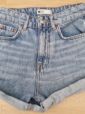 Blå denimshorts från Gina Tricot, storlek 30 - Klassiska blå denimshorts från Gina Tricot med hög midja och uppvikta benslut. Shortsen har fem fickor, bälteshällor och stängs med knapp och dragkedja. Perfekta för varma dagar och enkel att matcha med allt.