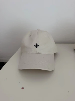 Morris keps  - Snygg beige keps med en liten svart broderad detalj framtill. Klassisk böjd skärm och justerbart spänne bak för perfekt passform. Tillverkad i bomullstyg som andas och passar till de flesta outfits.