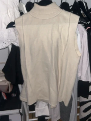 Beige ärmlös stickad polotröja - Säljer en beige ärmlös stickad polotröja med hög krage. Tröjan har en avslappnad passform och är gjord i mjukt stickat material. Perfekt att styla över en skjorta eller långärmad topp för en trendig lager-på-lager-look.