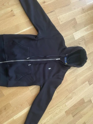 Svart hoodie från Polo Ralph Lauren - Svart hoodie med dragkedja från Polo Ralph Lauren. Klassisk design med vit broderad logga på bröstet, två fickor fram och justerbar huva med snören. Tillverkad i mjukt material som känns skönt mot huden. Perfekt för en avslappnad och stilren look.