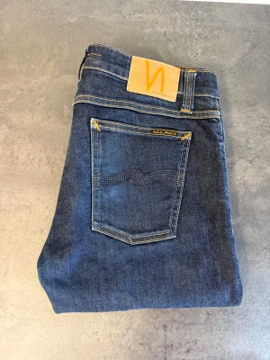 Mörkblå Nudie Jeans, Stl 33/32 - Snygga mörkblå jeans från Nudie Jeans med klassisk femficksmodell och gul kontrastsöm. Jeansen har en smal passform och är tillverkade i mjukt denimtyg. Perfekta för en stilren och avslappnad look. NYPRIS:2000kr