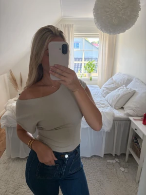 Beige oneshoulder topp - Snygg beige oneshoulder topp med tight passform. Toppen är croppad och har en enkel, stilren design utan mönster eller detaljer. Perfekt att matcha med jeans eller kjol för en trendig look.