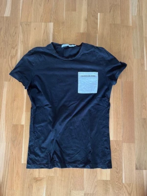 Svart t-shirt med ficka Calvin Klein medium - Svart t-shirt från Calvin Klein med ljusgrå bröstficka där det står 'Calvin Klein Jeans'. Klassisk rund hals och korta ärmar. Perfekt till jeans eller shorts för en clean och enkel look. Medium