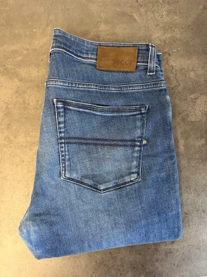 Boggi Milano Claude Jeans, stl 34 - Säljer ett par klassiska blå jeans från Boggi med rak passform och fem fickor. Jeansen har snygga slitningar och kontrastsömmar samt en brun läderpatch med logga bak i midjan. Perfekta till vardagslooken! NYPRIS: 2500kr