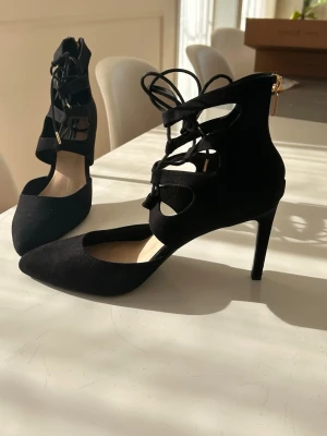 Svarta spetsiga pumps med snörning - Snygga svarta pumps med spetsig tå och höga klackar. Skorna har dekorativ snörning framtill och dragkedja bak för enkel påtagning. Materialet ser ut som mocka och designen har öppna partier på sidorna.