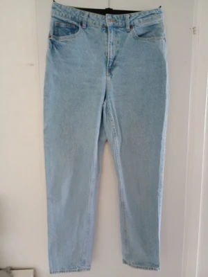 Ljusblå mom jeans med hög midja - Säljer ett par ljusblå mom jeans i klassisk modell med hög midja och raka ben. Jeansen har fem fickor och bälteshällor. Tillverkade i ett kraftigt denimtyg som ger en retrovibe. Perfekta för dig som gillar en avslappnad och trendig stil.