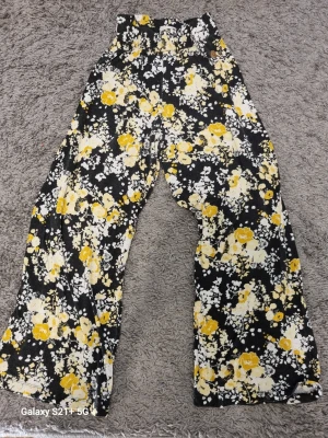 Blommiga vida leggings i svart och gult - Säljer ett par snygga leggings med svart bas och blommigt mönster i gult och vitt. Modellen är högmidjad och har vida ben för en avslappnad look. Perfekta för dig som vill sticka ut med färg och mönster. Materialet känns mjukt och stretchigt.