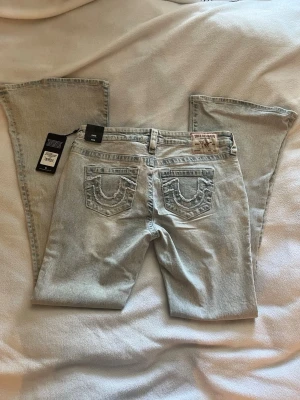 Ljusblå bootcut jeans True Religion - Säljer ett par ljusblå bootcut jeans från True Religion med ikoniska broderade bakfickor och patch med logga bak i midjan.💫 Jeansen har klassisk femficksdesign och är tillverkade i denim med lätt tvättad look. Perfekta för dig som gillar street och trendig stil. 🔥 Modellen heter: CARRIE low rise flare 🌟Storlek: W29, Innerbenslängd: 86cm 