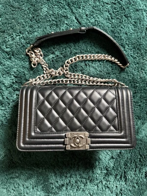 Svart Boy Bag  - Elegant svart quiltad väska tillverkad av syntetmaterial med gulddetaljer och kedja. Har dragsko och kedjestängning, samt fram- och sidfickor med märkeslogotyp. Perfekt storlek för att bära det viktigaste och ge din look en touch av lyx.
