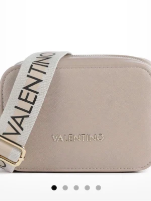 Beige axelväska från Valentino - Snygg beige axelväska från Valentino med justerbar bred axelrem i textil med logga. Väskan har en stilren, boxig form och är gjord i ett slätt material med guldfärgade detaljer. Perfekt för dig som vill ha en trendig och praktisk väska.